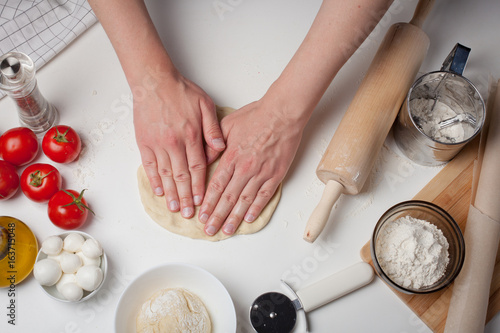 Male hands preparing dough for pizza on table closeup. On the white table are the tomatoes, mozzarella balls, olive oil and flour