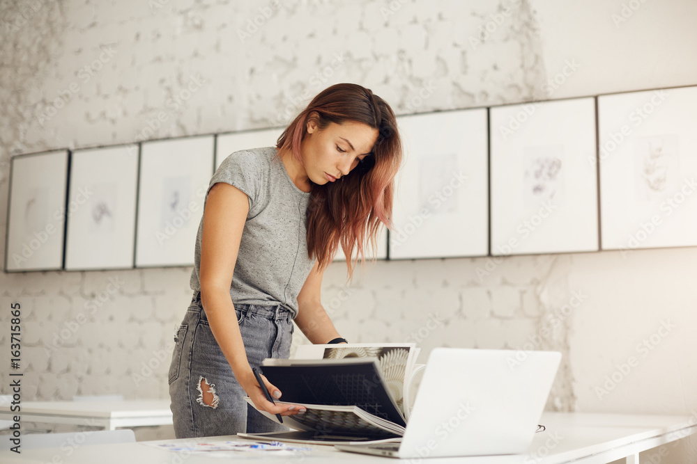 Young female clothes designer browsing through magazine using laptop computer to find trending textiles. Young female clothes designer browsing through magazine using laptop computer to find trending textiles.