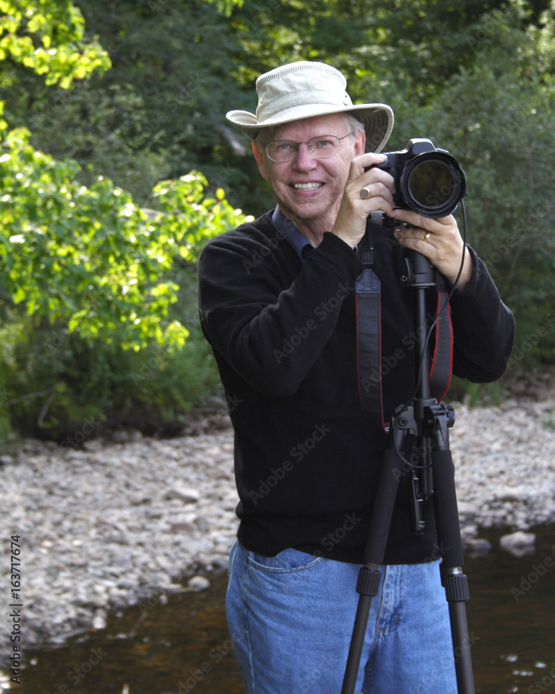 a senior photographer poses with his camera and hat outside smiling