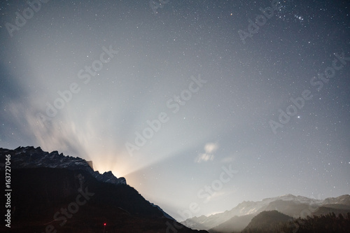 Moon rising with ray light behide the mountain with little snow on the top and moving cloud in the blue color night with stars in winter at Lachung in North Sikkim, India. © artitwpd