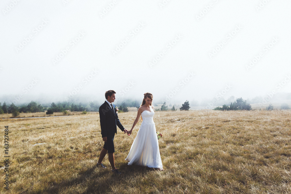 Young bride and groom walking high in mountains. Amazing weather with fog and sun Young bride and groom walking high in mountains. Amazing weather with fog and sun