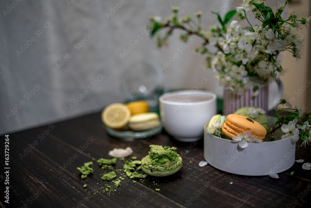 Crushed pastel, orange and green macaroons on a wood background with whole one in white plate with jug with flowers on the back Crushed pastel, orange and green macaroons on a wood background with whole one in white plate with jug with flowers on the back