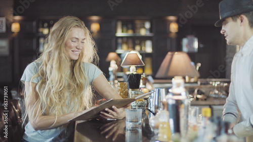 Bartender helps to blonde 20s woman make a choice in menu of bar