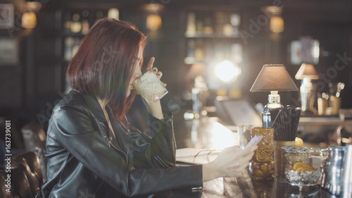 20s sexy beauty redhead woman in black leather jacket is drink cocktail, looking at camera and flirt smiles in the bar