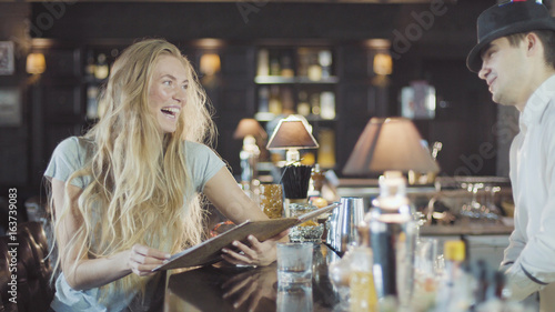 Bartender helps to blonde 20s woman make a choice in menu of bar