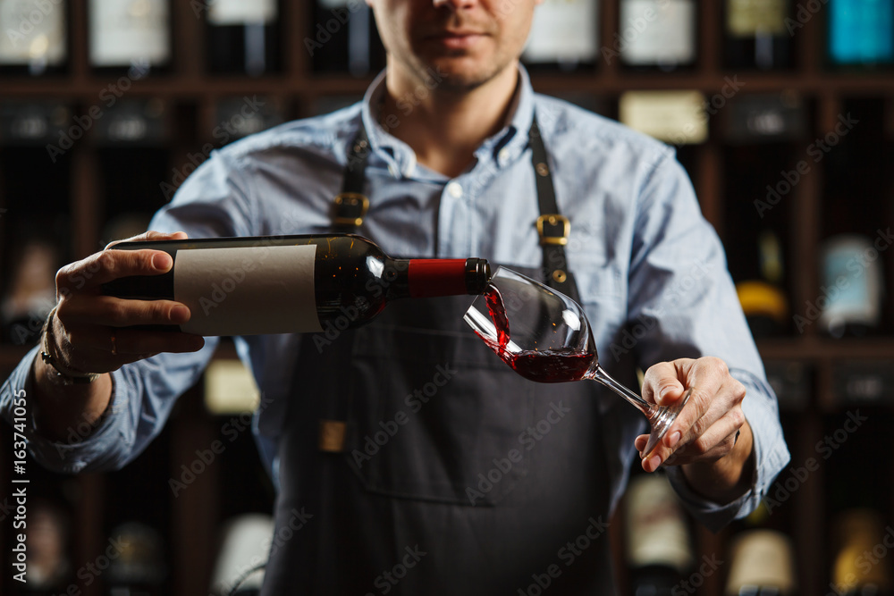 Male sommelier pouring red wine into long-stemmed wineglasses. - obrazy, fototapety, plakaty 
