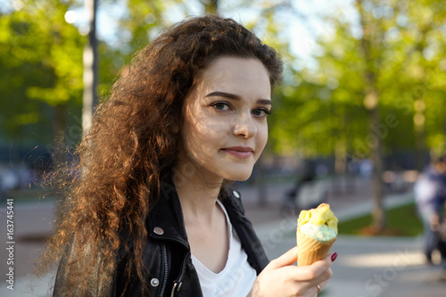 Headshot of pretty girl standing outdoors holding waffle cone  eating delicious fruit ice-cream during walk in city park  looking at camera with cute charming smile. People and lifestyle concept