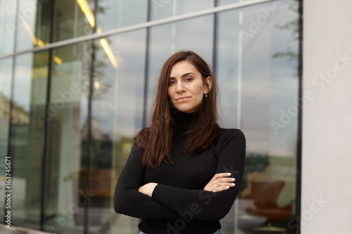 Urban street portrait of serious beautiful young European woman with long dark hair posing against glass windows office building, keeping arms crossed and looking at camera with confident expression Free Download Urban street portrait of serious beautiful young European woman with long dark hair posing against glass windows office building, keeping arms crossed and looking at camera with confident expression - GFXLoad