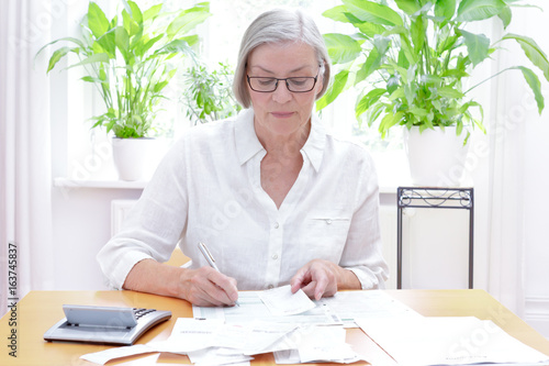 Deutsche Seniorin füllt die Formulare für die jährliche Steuererklärung aus, im Wohnzimmer an einem Tisch mit Taschenrechner und vielen Rechnungen und Quittungen. photo