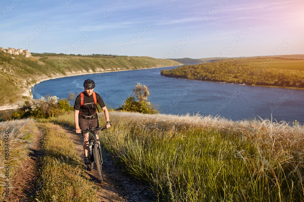 Portrait of the young cyclist standing on the hill above the river against blue sky with clouds.