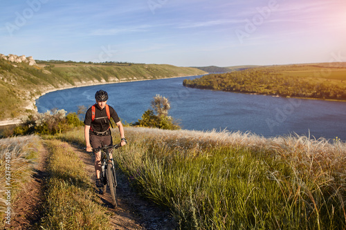 Portrait of the young cyclist standing on the hill above the river against blue sky with clouds.