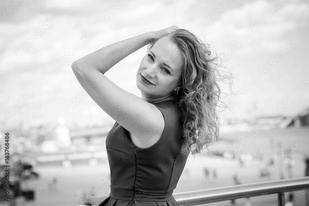 Portrait of blonde hair curly woman with healthy skin wearing blue dress looking at camera with  pensive expression. model with fair hair posing outdoors in city, architecture background