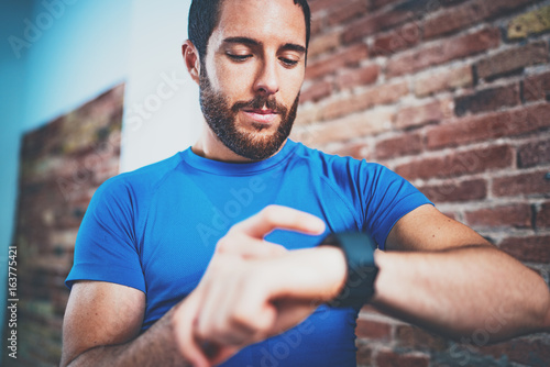 Young Muscular bearded athlete checking burned calories on electronic smart watch application after good indoor workout session in fitness gym.Brick wall on blurred background.