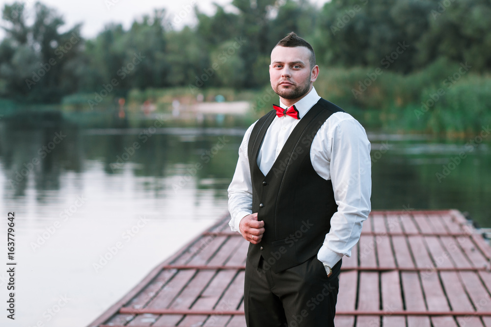 Young stylish man in a waistcoat, horizontal portrait of the groom, portrait on a background of nature, the river and the pier, look at the camera Young stylish man in a waistcoat, horizontal portrait of the groom, portrait on a background of nature, the river and the pier, look at the camera