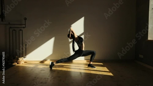 The athetic girl is standing in the pose of lunges, holding the neck and moving back near the window. photo
