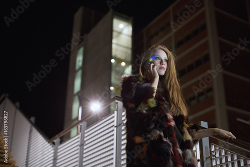 Cool young woman with colorfull fur and mobile phone going out at night walking in city street photo