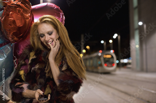 Beautiful young woman with multi colored fur and balloons smiling on train platform at night photo