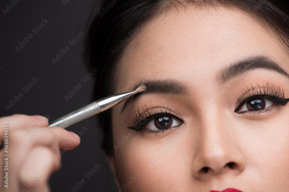 Close up of beautiful face of young asian woman getting make-up. The artist is applying eyeshadow on her eyebrow with brush