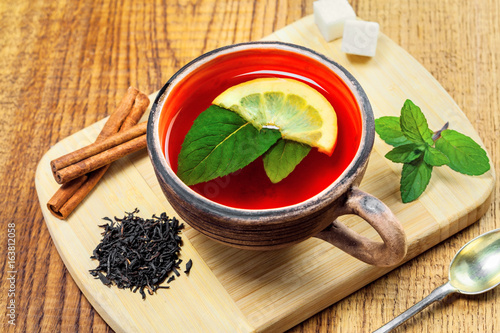 Hot drink in a rustic clay cup made of black tea, fresh mint leaves and lemon on a wooden table. Delicious healthy beverage made of herbals and fruits.