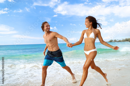 Happy beach vacation couple having fun running splashing water at tropical travel vacations.
