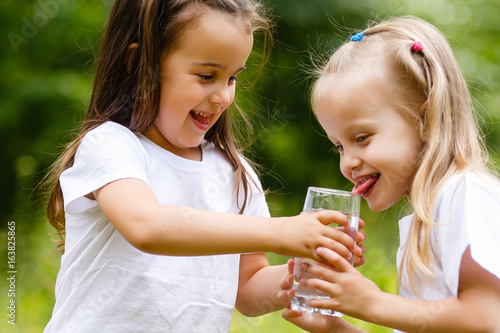 Two little girls are drinking a glass of water in the park. Concept of purity, ecological and biological product, love for nature