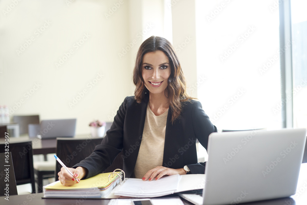 Working in the office. Shot of a happy young businesswoman sitting at desk in front of laptop and doing some paperwork. Working in the office. Shot of a happy young businesswoman sitting at desk in front of laptop and doing some paperwork.
