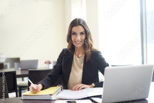 Working in the office. Shot of a happy young businesswoman sitting at desk in front of laptop and doing some paperwork.  © gzorgz
