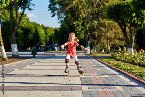 Pretty little girl in red t-shirt learning to roller skate outdoors on beautiful summer day