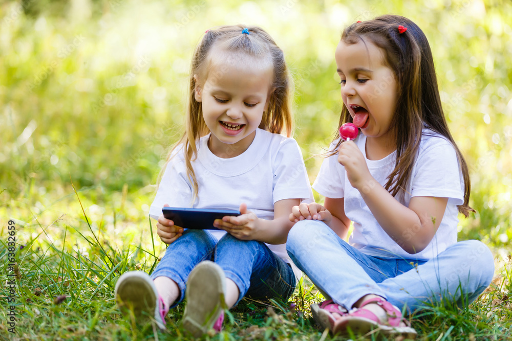 Two small laughing girls sit together at the outdoor in the city and play with smartphone Two small laughing girls sit together at the outdoor in the city and play with smartphone