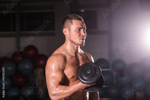 Handsome power athletic guy bodybuilder doing exercises with dumbbell. Fitness muscular body on dark background