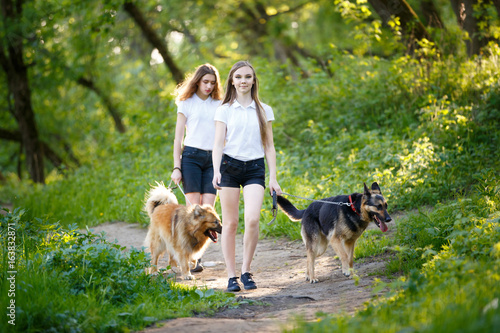 Two smiling teenage girls walking with her dogs in spring park. Friendship concept background
