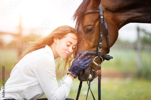Young smiling rider woman in white shirt leaning to horse head. Friendship equine concept background