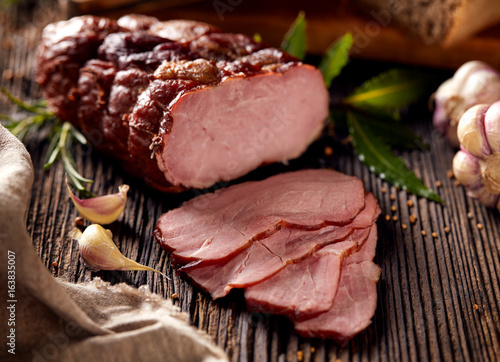 Smoked ham sliced on a wooden rustic table with addition of fresh aromatic herbs.  Natural product from organic farm, produced by traditional methods