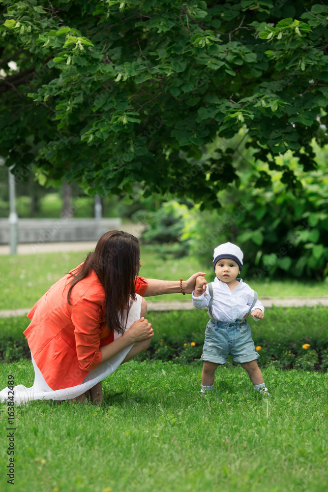A small boy with dark eyes in a white shirt and denim shorts, trying to take the first steps with the help of his mother