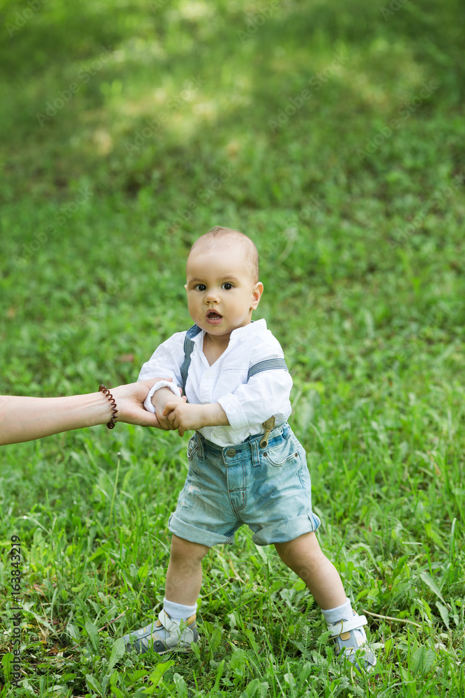 A little boy with dark eyes in a white shirt and denim shorts, trying to make the first steps on the green grass A little boy with dark eyes in a white shirt and denim shorts, trying to make the first steps on the green grass