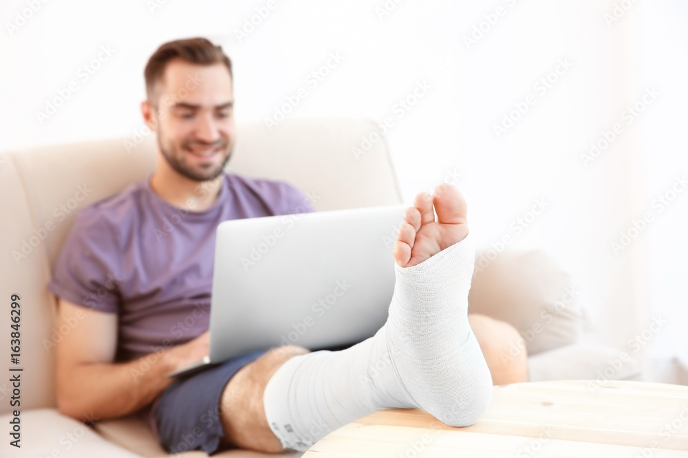 Handsome young man with laptop and bandaged leg sitting on sofa at home Handsome young man with laptop and bandaged leg sitting on sofa at home