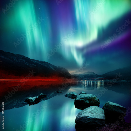 A large Northern Lights (aurora borealis) display glowing over a mountain pass and reflected on a lake at night. Photo composition.