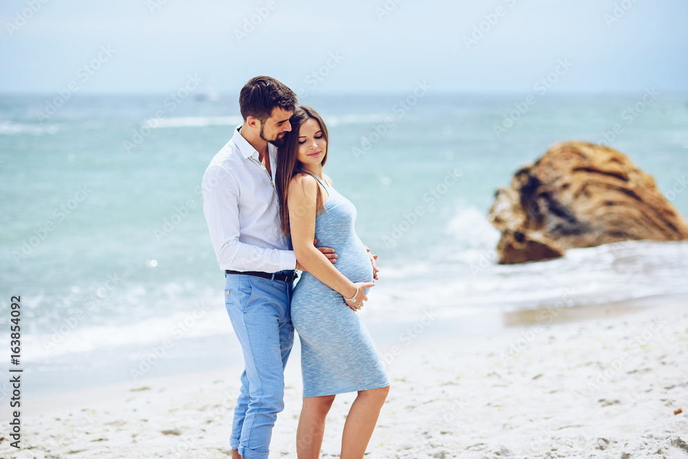 Courageous and handsome man in a white shirt and blue trousers hugs her beautiful pregnant wife in a blue dress against the background of the rocks and the sea. Courageous and handsome man in a white shirt and blue trousers hugs her beautiful pregnant wife in a blue dress against the background of the rocks and the sea.