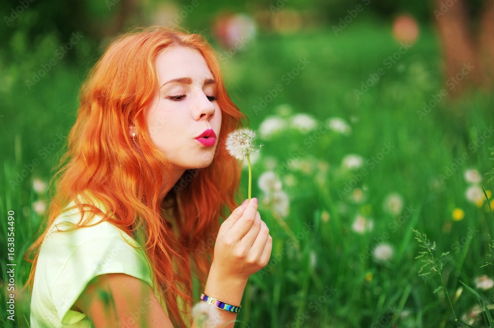Young beautiful red-haired girl sitting on green grass in a field, holding flower a dandelion and blowing on it. Closeup portrait. Young beautiful red-haired girl sitting on green grass in a field, holding flower a dandelion and blowing on it. Closeup portrait.