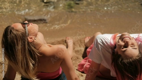 Two women sitting on beach in warm weather. Located on coastine leaning their hands on sand, they recline back and put their face under rays of tender sun, enjoying it. Attractive ladies wearing photo