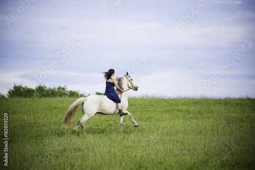 horizontal girl riding white horse bareback across meadow with big sky and green grass