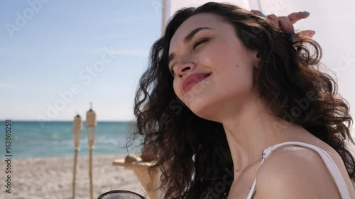 portrait girl with curls, women smiles and drinks cocktail from oconut, wind develops female hair, summer season, expensive vacation, background sea ocean and sand, tropical islands photo