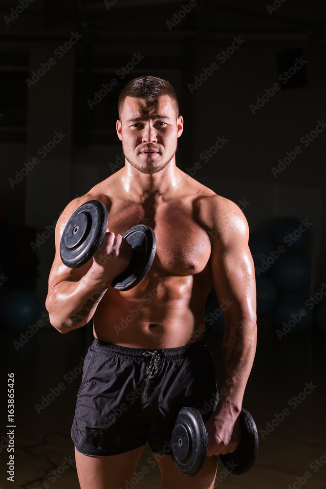 Athletic shirtless young male fitness model holds the dumbbells.
