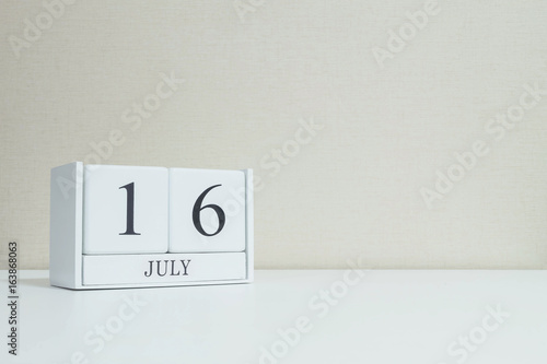 Closeup white wooden calendar with black 16 july word on blurred white wood desk and cream color wallpaper in room textured background with copy space , selective focus at the calendar