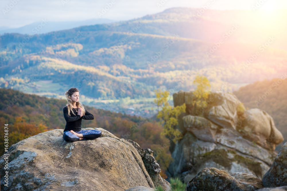 Pretty young woman is practicing yoga and doing asana Siddhasana on the top of the mountain in the evening. Autumn forests, rocks and hills on the background