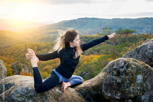 Flexible young girl is practicing yoga and doing asana Eka Pada Rajakapotanasana on the top of the huge boulder in the evening. Beautiful sunset, autumn forests, rocks and hills on the background