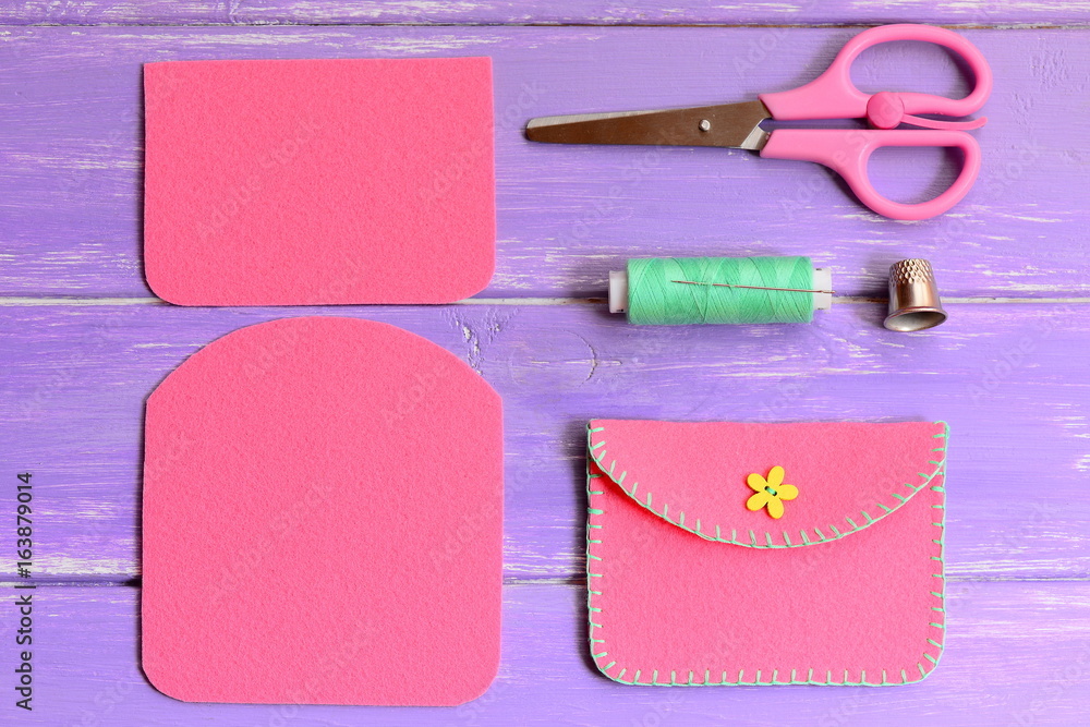 Obraz premium Children pink felt purse with yellow flower wooden button. Scissors, thread, needle, thimble on a wooden table. Easy homemade DIY made of felt. Top view