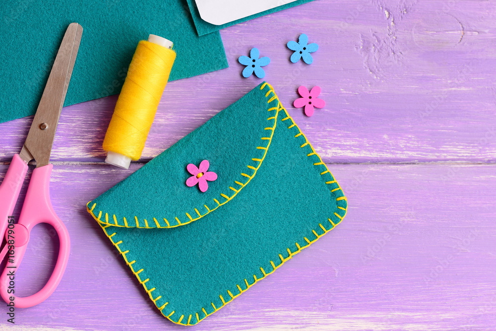 Naklejka premium Small felt purse with flower wooden button. Scissors, thread, flower wooden button on a wooden background with copy space for text. Hand sewing for kids