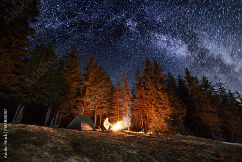 Male tourist have a rest in his camp near the forest at night. Man sitting near campfire and tent under beautiful night sky full of stars and milky way, and enjoying night scene