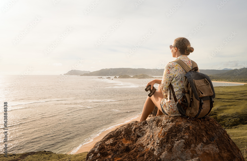 © soft_light - Woman traveller with backpack and binoculars in hands enjoying view coast on sunset